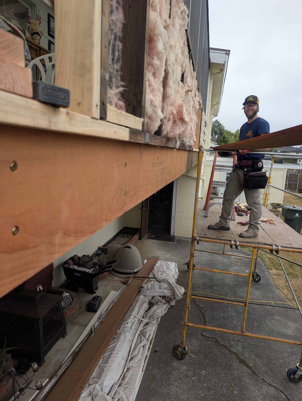 Smiling crew member in branded NorCal Exterior cap on scaffolding during the same repair project, fresh framing and insulation visible behind